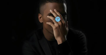 Male model wearing a unique blue mechanical ring watch, hand near face, black background.