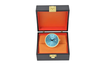 Eine elegante, runde Tischuhr mit hellem blauen Zifferblatt und goldfarbenem Lunette ruht in einer stilvollen schwarzen Präsentationsbox, die mit lebendigem orangefarbenem Samt ausgekleidet ist.